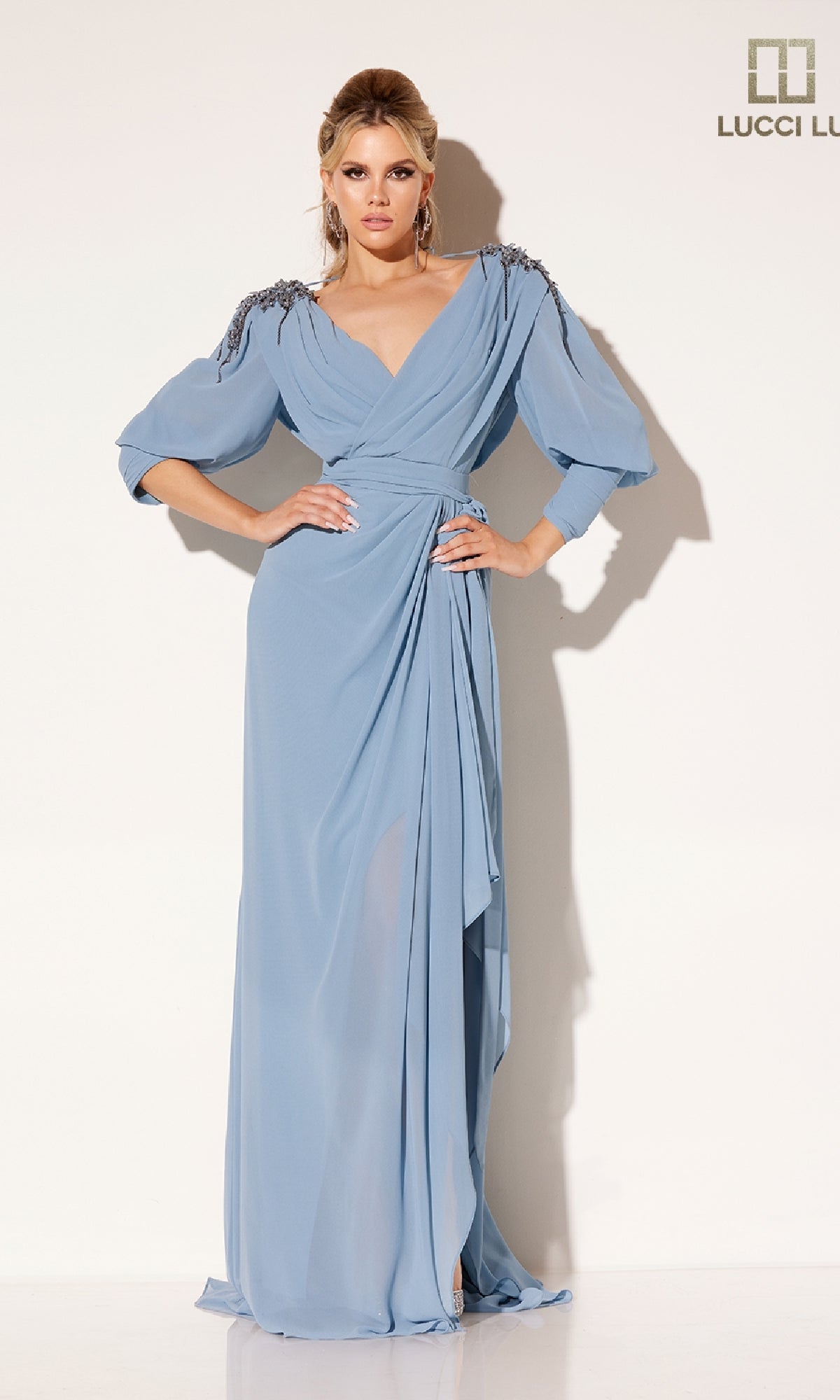 A woman poses in the Lucci Lu C8092 long formal dress, featuring floor-length draped light blue fabric, embellished shoulders, and long sleeves, set against a plain white background.